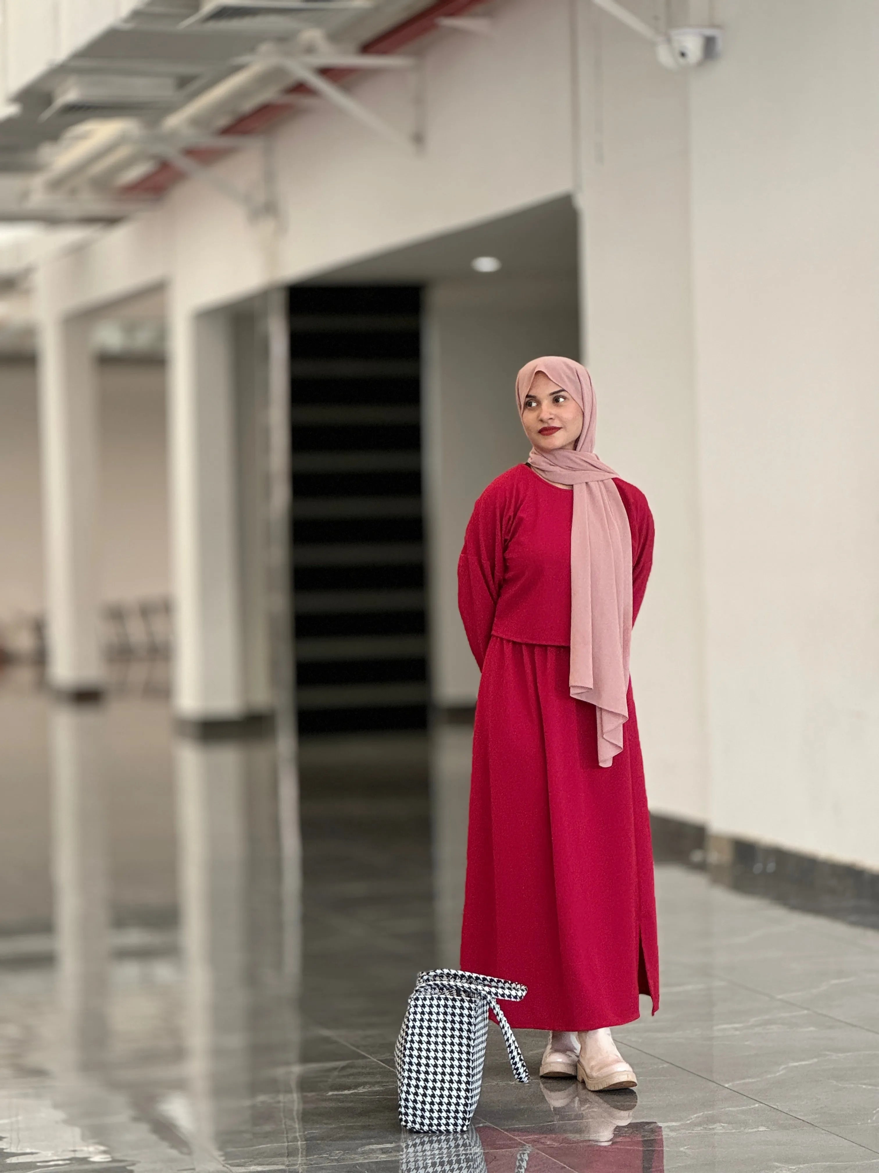 Model wearing a reddish maroon top and skirt with a matching headscarf in a modern indoor setting