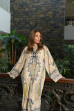 Woman wearing a beige printed modest kaftan dress with intricate blue patterns standing near an ornate railing indoors.