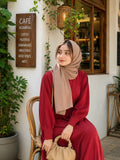 Woman in a maroon modest maxi dress and beige hijab sitting on a wooden chair outside a café holding a beige handbag.