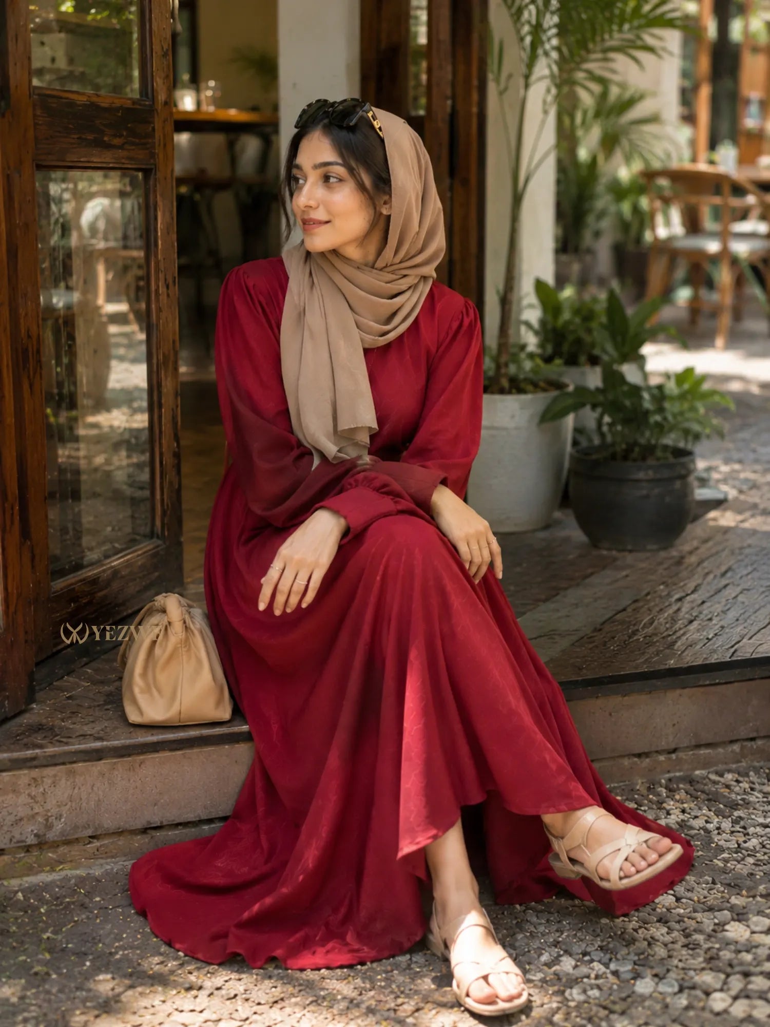 Woman sitting outdoors wearing a maroon modest maxi dress with long sleeves and beige hijab near a café entrance.