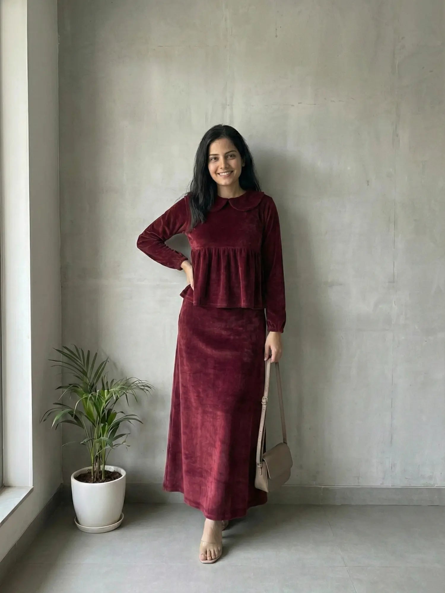 woman wearing wine maroon velvet modest maxi dress standing indoors in a stylish pose with hand on waist and carrying a crossbody bag