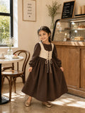 Junior girl wearing a brown cottagecore maxi dress with cream corset-style bodice and puff sleeves in a warm café interior.