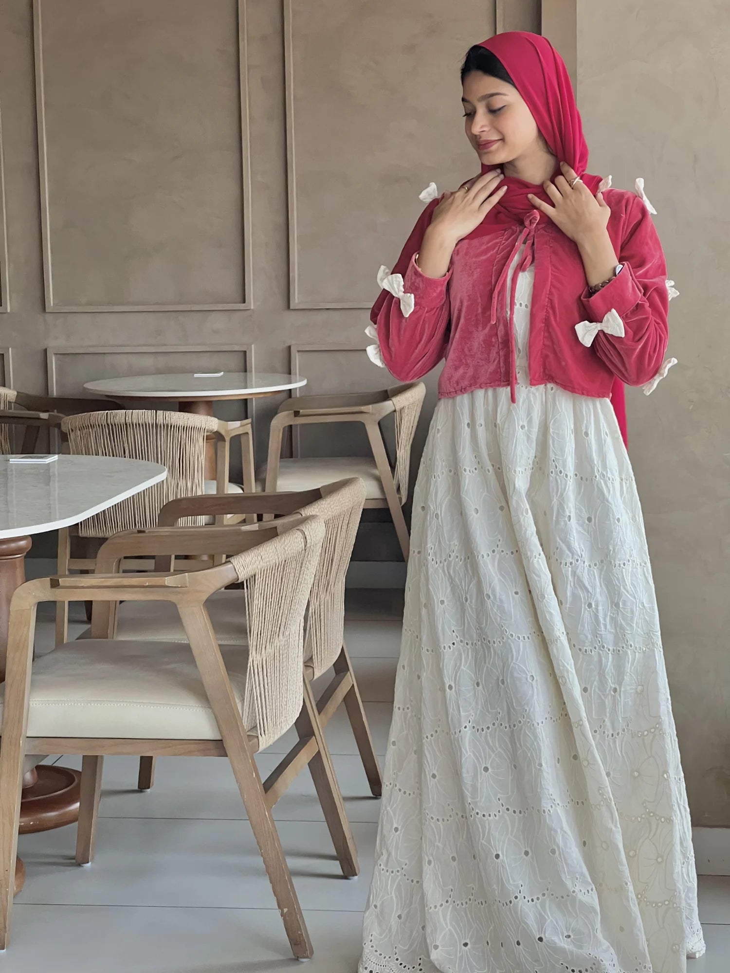 A woman wearing a red hijab and a modest outfit with a maroon top and a long white embroidered maxi skirt, standing indoors and adjusting her scarf.