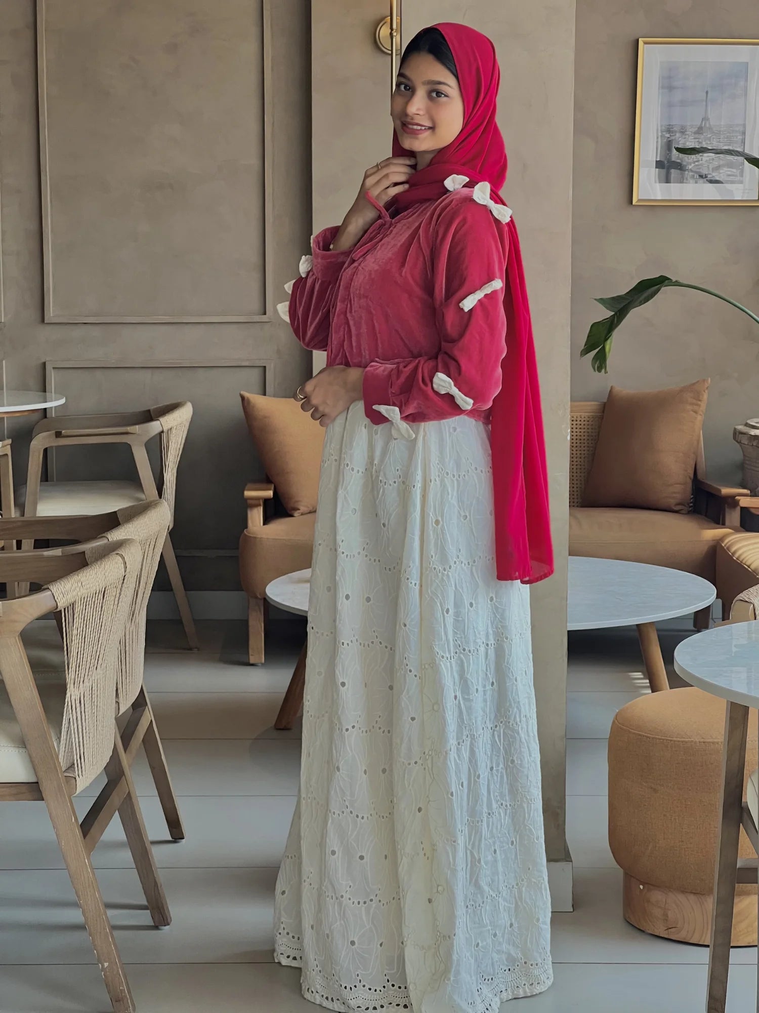 A woman wearing a red hijab with a modest vintage-style outfit, featuring a maroon top and a long white embroidered maxi skirt, standing in a cozy indoor setting with wooden furniture.