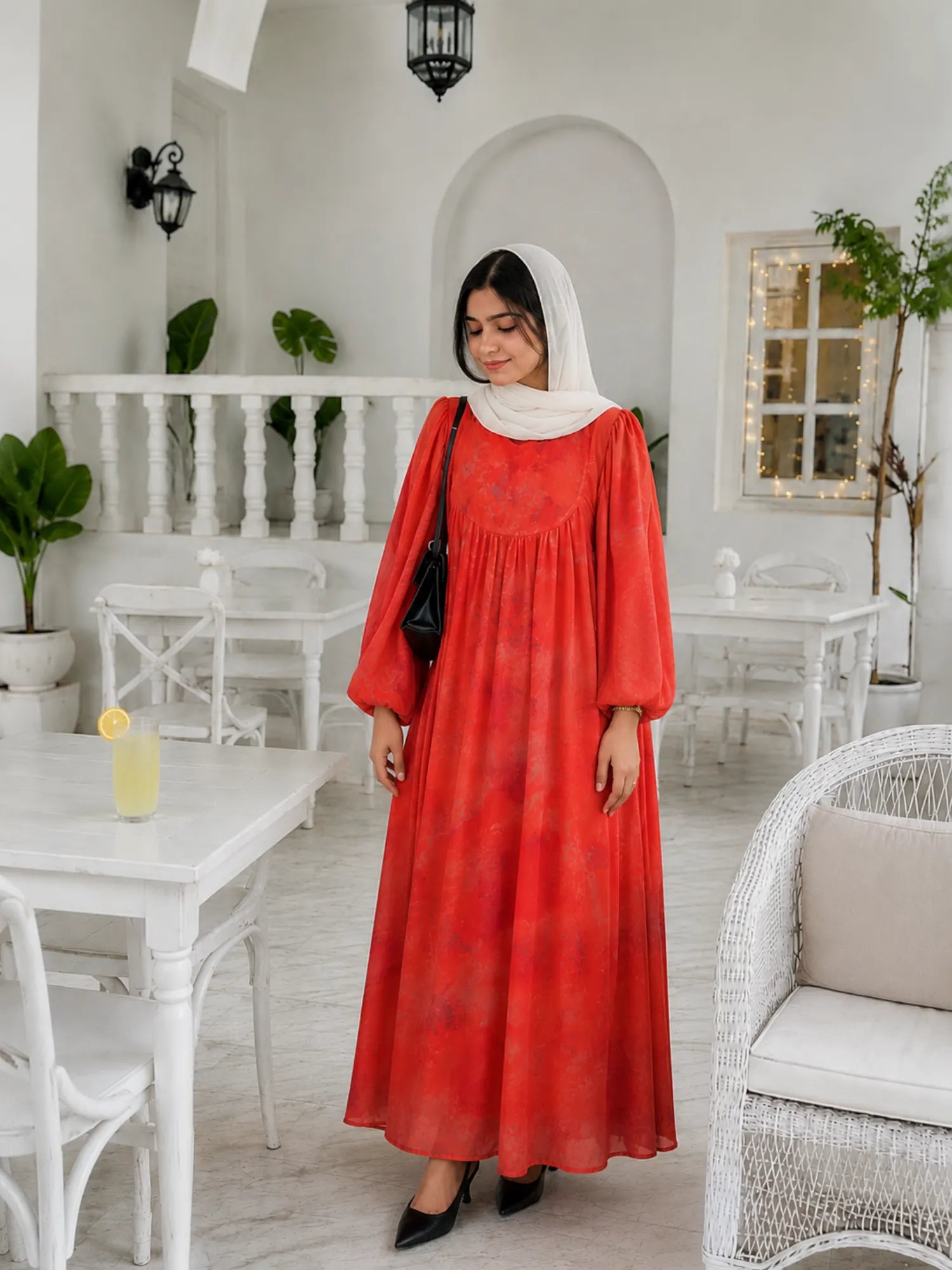 Woman wearing a coral red modest maxi dress with puff sleeves and white hijab standing in a bright elegant café interior.