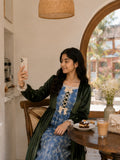 Junior girl wearing a modest green velvet and blue patterned maxi dress, sitting in a cozy café while taking a selfie with her phone.