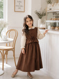 Little girl wearing a brown maxi dress with cream puff sleeves, posing near a bakery counter in a vintage style party wear look