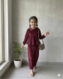 Young girl wearing a maroon cotton co-ord set with long sleeves and matching pants, paired with a small sling bag, standing in a minimal indoor setting.