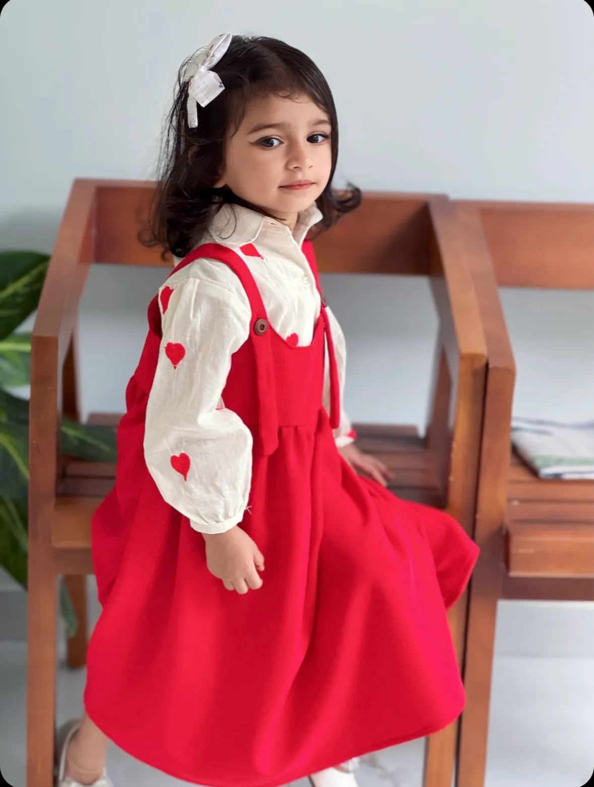 Young girl wearing a red pinafore for junior girls over a white blouse sitting on a wooden bench