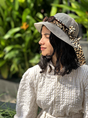 Model wearing a white textured jumpsuit with a stylish hat and leopard print ribbon, surrounded by greenery