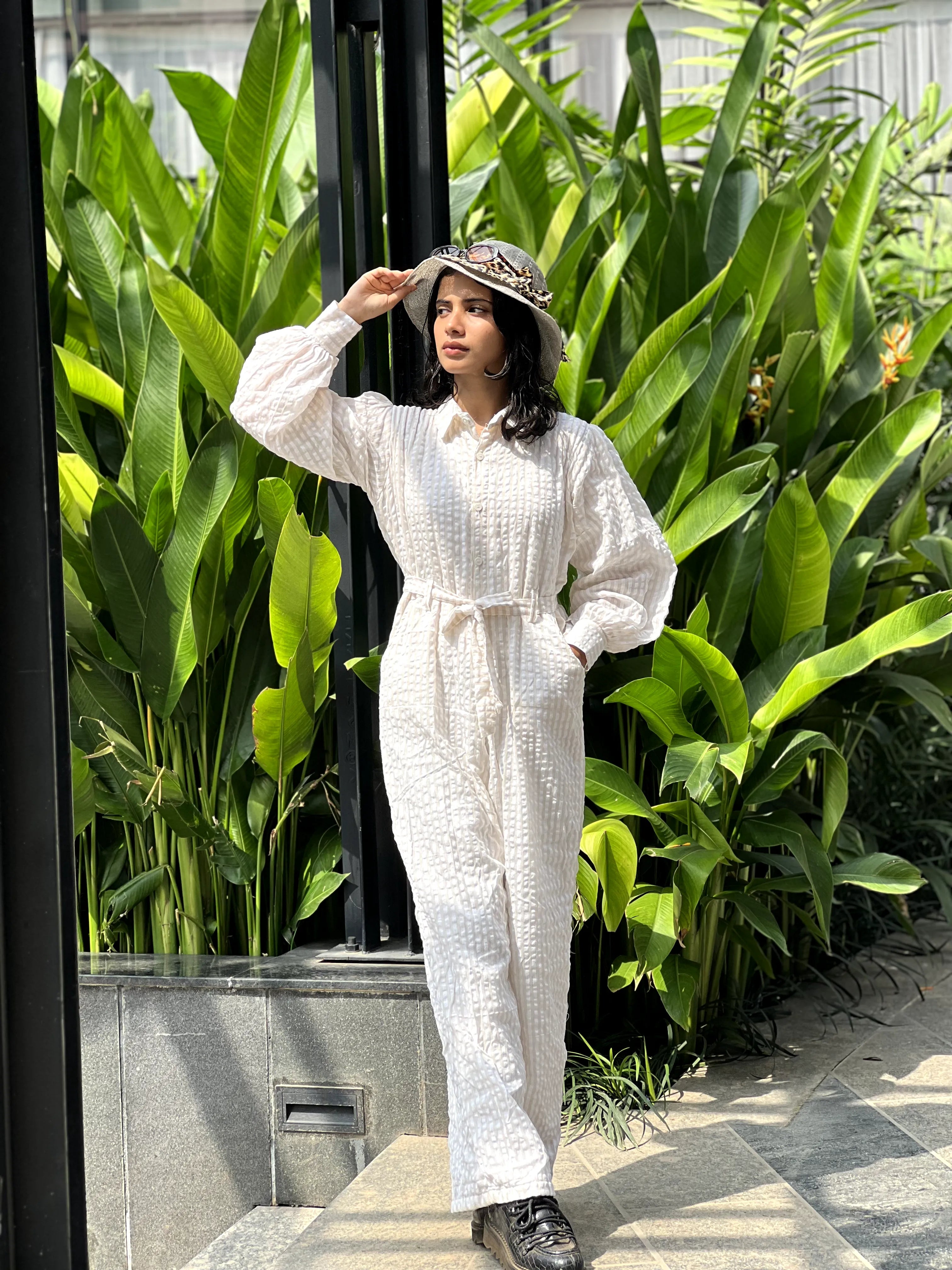 Model wearing a stylish white textured jumpsuit with a belted waist in a tropical setting