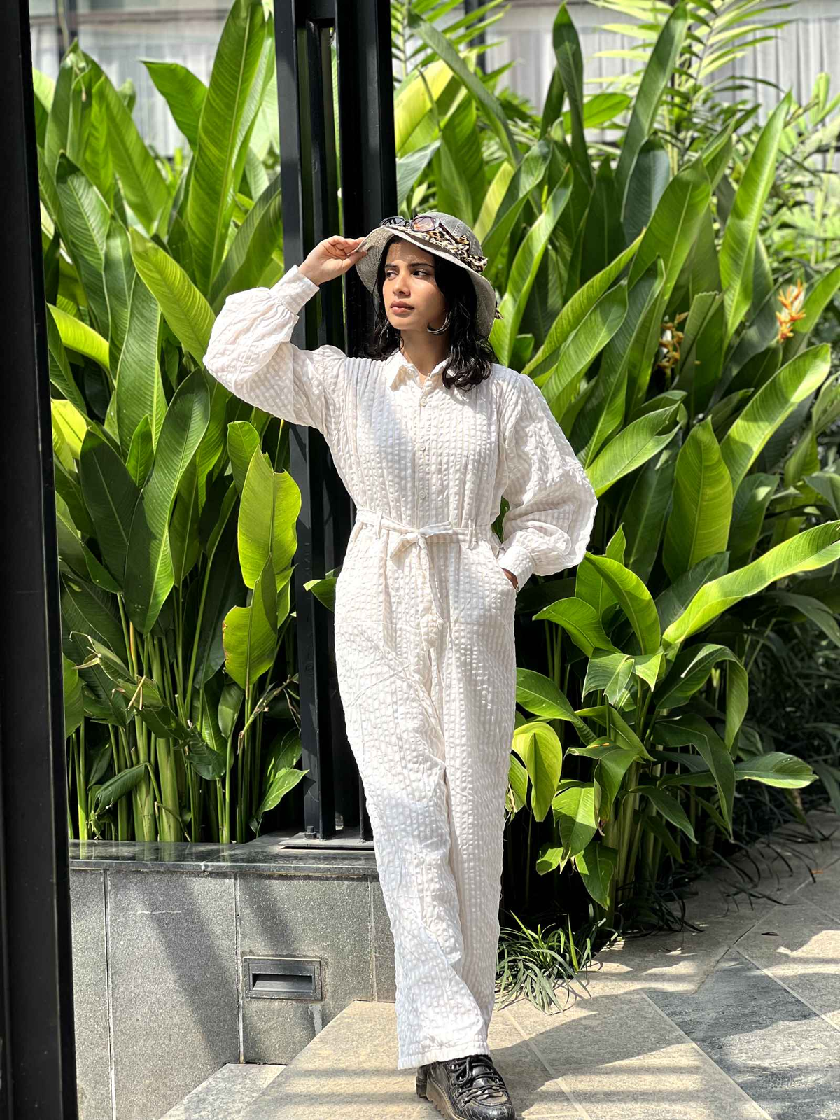 Model wearing a stylish white textured jumpsuit with a belted waist in a tropical setting