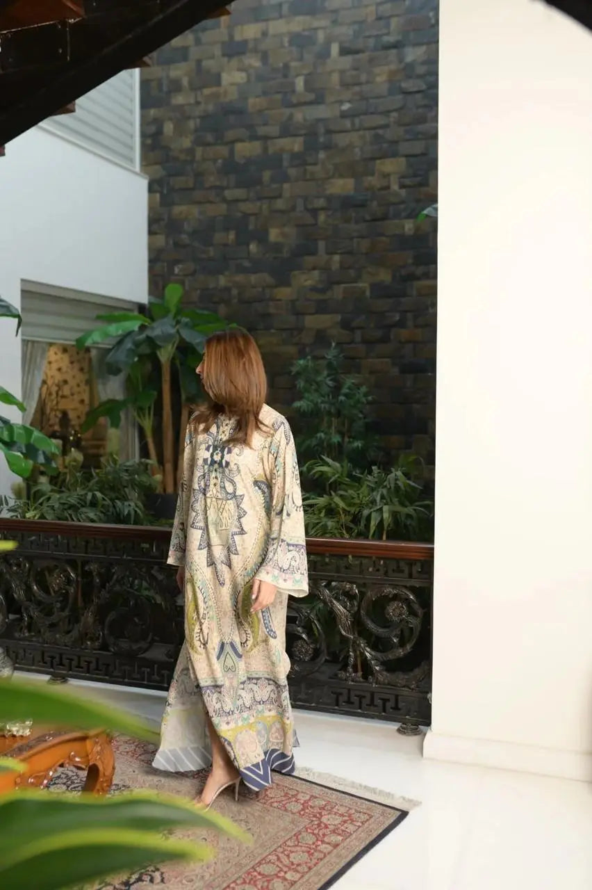 Back view of a woman wearing a beige paisley print maxi dress with blue detailing standing beside an ornate railing in an elegant indoor setting.