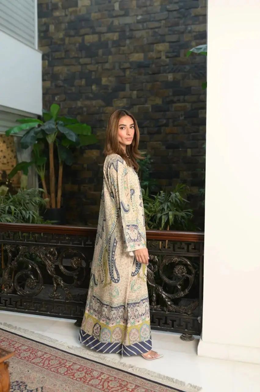 Woman wearing a beige paisley print maxi dress with blue and pastel detailing standing indoors beside a decorative black railing.