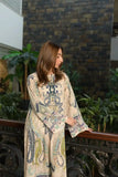 Woman wearing a beige printed kaftan maxi dress with blue and pastel paisley patterns standing beside an ornate railing indoors.