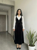 woman wearing black velvet pinafore modest maxi dress with white shirt standing indoors in full front view