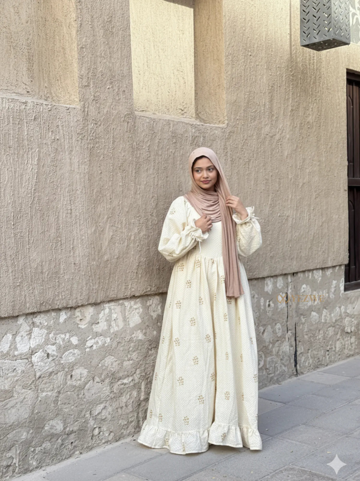 young woman wearing cream embroidered modest maxi dress with hijab standing outdoors near beige wall full length view
