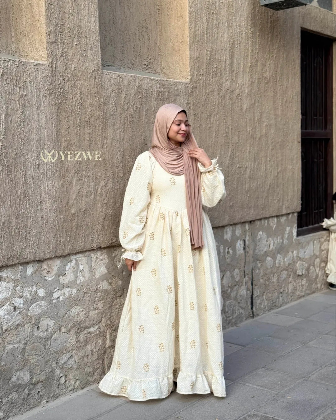 young woman wearing cream embroidered modest maxi dress with hijab standing side pose outdoor street view
