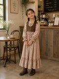 Young girl wearing a beige floral Linen Lux maxi dress with brown corduroy bodice and long sleeves standing in a warm café setting.