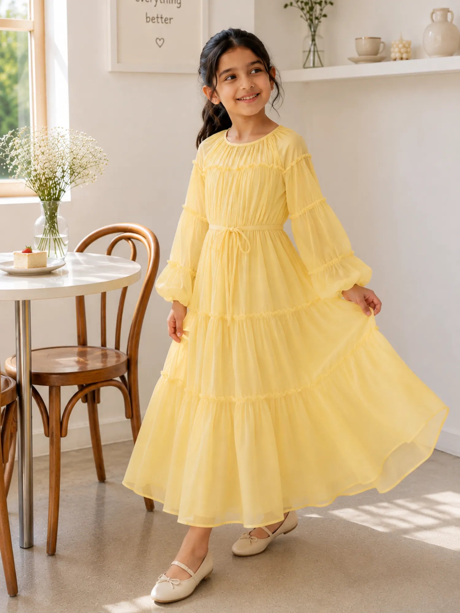 Young girl twirling in a pastel yellow flowy tiered maxi dress with long puff sleeves in a bright indoor café-style setting.