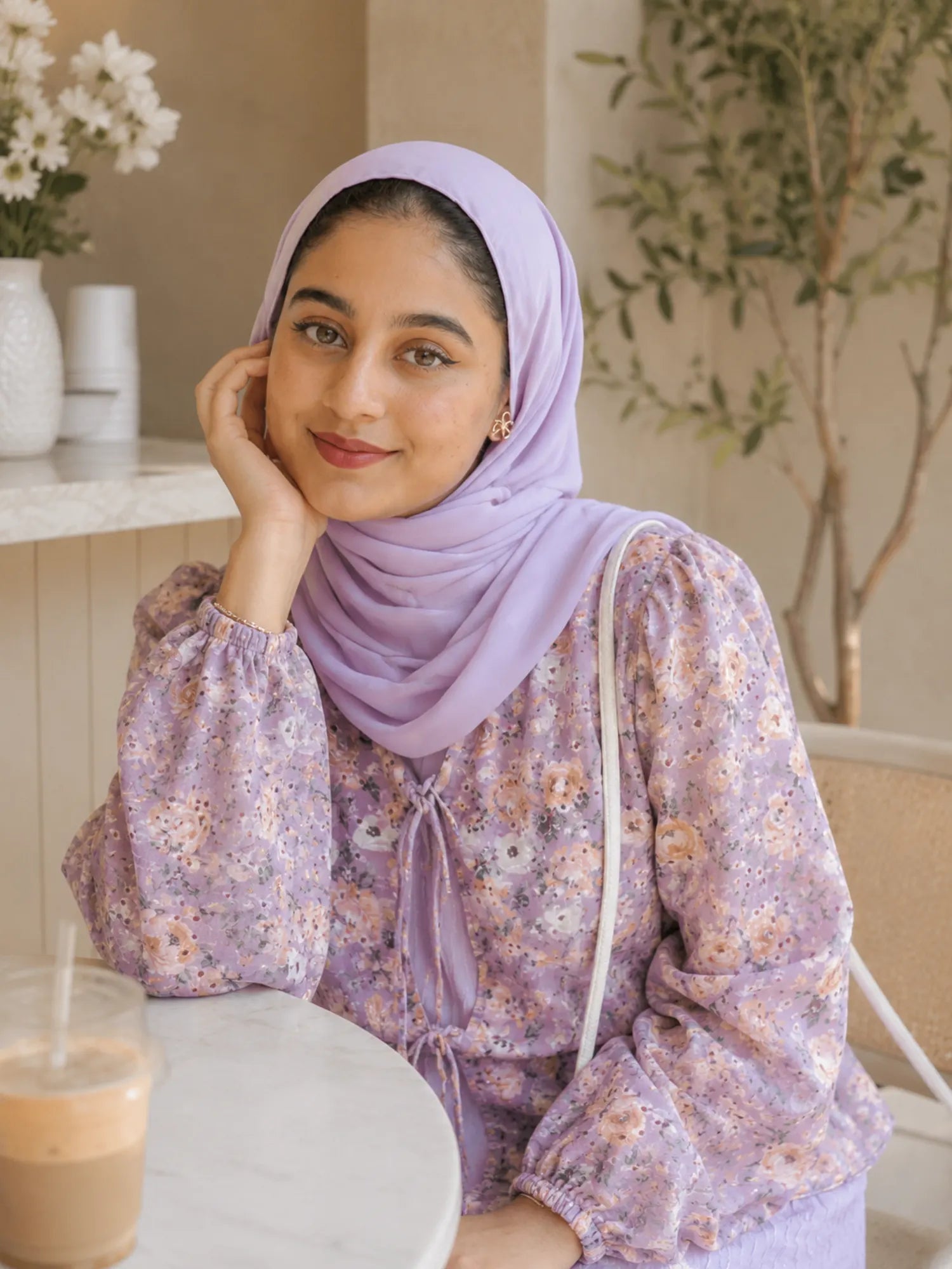 Close-up portrait of a woman wearing a lavender floral modest dress with matching hijab sitting at a café table.