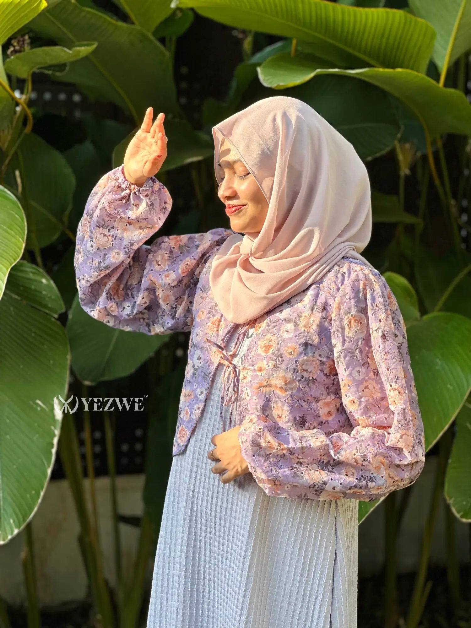 Woman wearing a lavender floral modest dress with beige hijab standing outdoors near large green leaves in sunlight.