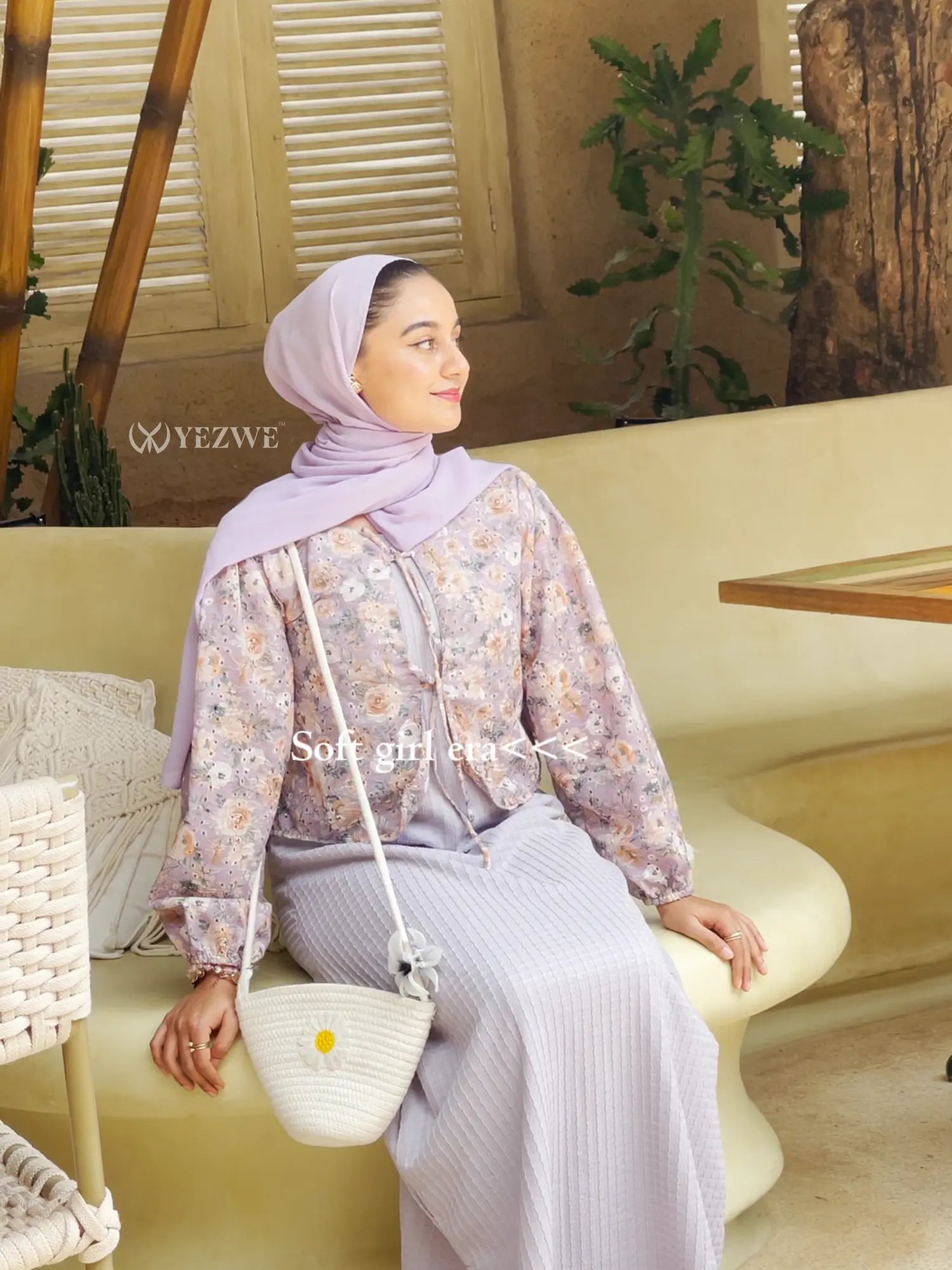 Woman wearing a lavender floral modest maxi dress with matching hijab, sitting on a cream sofa with a white woven handbag in a cozy indoor setting.