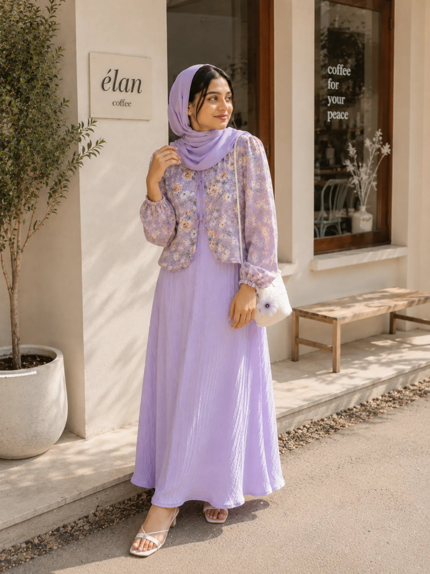 Woman wearing a lavender floral modest maxi dress with matching hijab standing outside a café with a white handbag.
