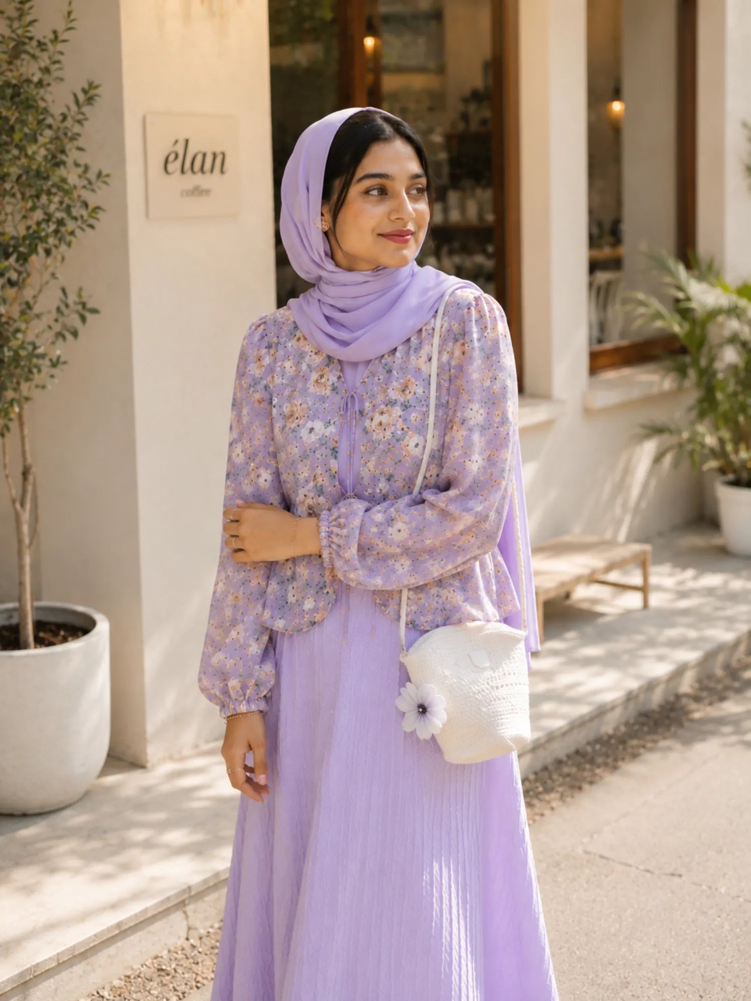 Woman wearing a lavender floral modest maxi dress with matching hijab and white handbag standing outside a café in soft daylight.