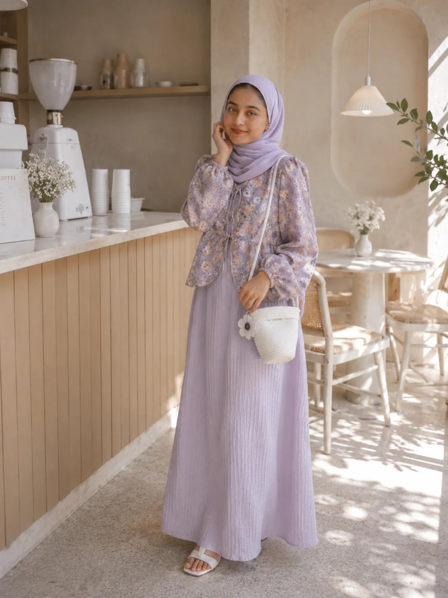 Woman wearing a lavender modest maxi dress with floral top and matching hijab, standing in a soft café interior with a white handbag.