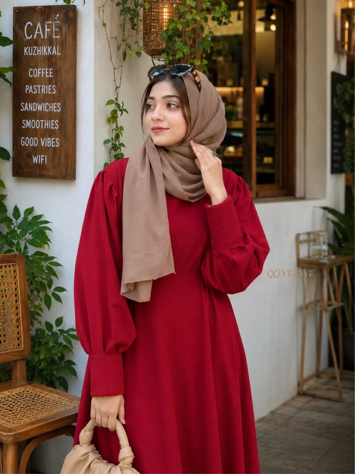 Woman wearing a maroon modest maxi dress with puff sleeves and beige hijab standing near a café with a handbag.