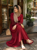 Woman sitting outdoors wearing a maroon modest maxi dress with long sleeves and beige hijab near a café entrance.