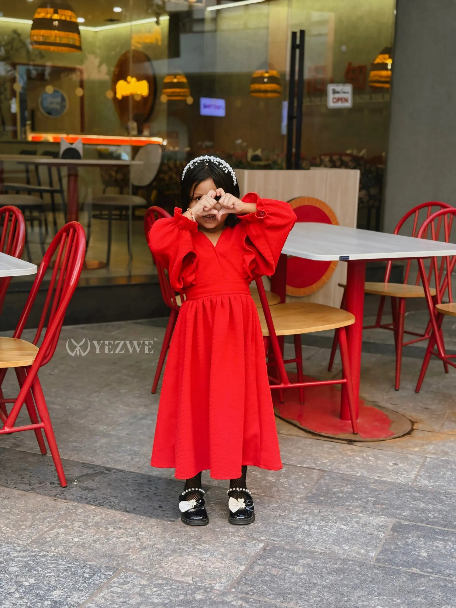 Junior girl wearing a bright red maxi dress for junior girls with puffed sleeves and black shoes in a cafe setting
