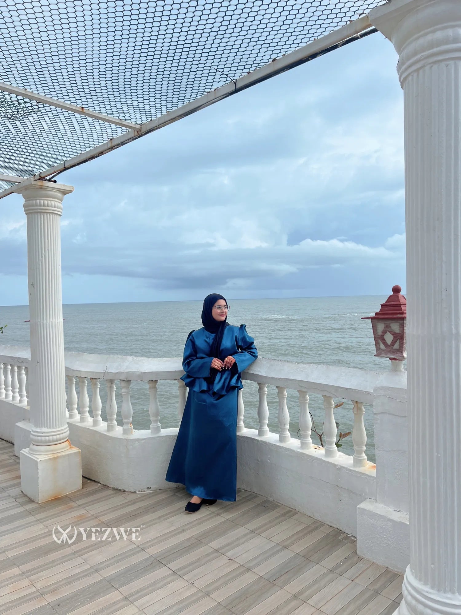 Woman wearing a blue modest co-ord set by YEZWE standing on a balcony overlooking the ocean