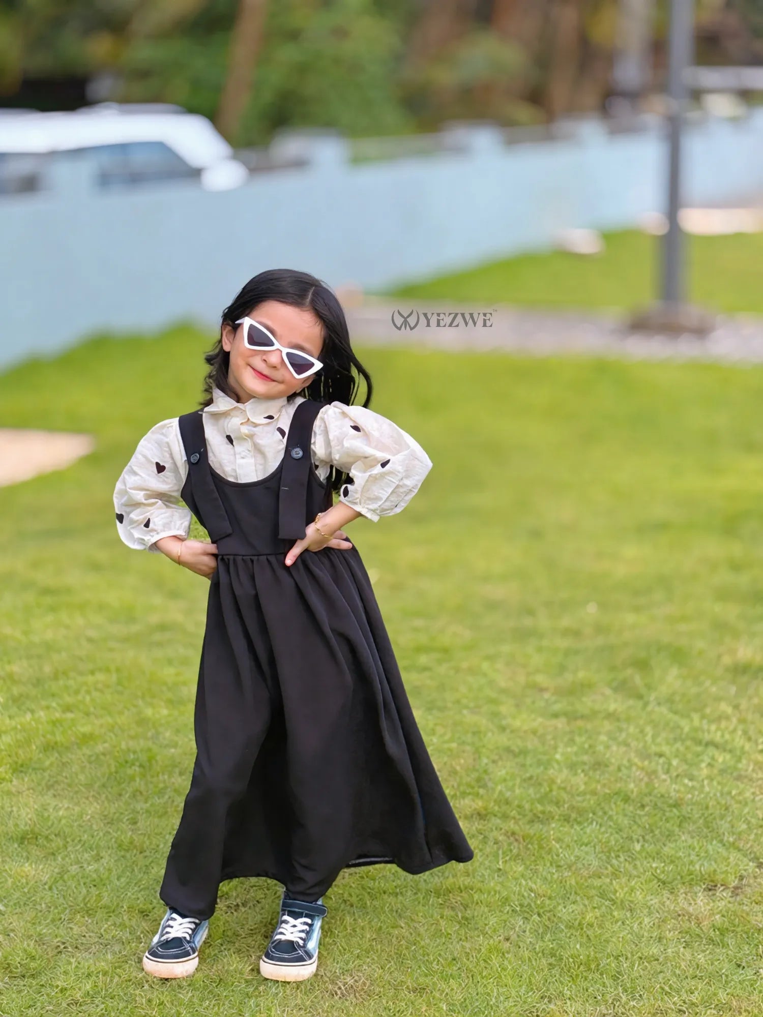 YEZWE junior girls Pinafore Dress black pinafore layered over printed blouse styled with white sunglasses and sneakers
